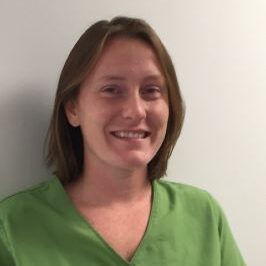 A person with shoulder-length hair smiling, wearing a green top, standing against a plain light-colored background.