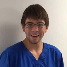 Man wearing glasses and blue scrubs, smiling in front of a plain background.