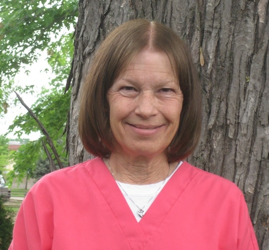 A woman with shoulder-length brown hair is wearing a pink top and smiling, standing in front of a tree trunk with green leaves.