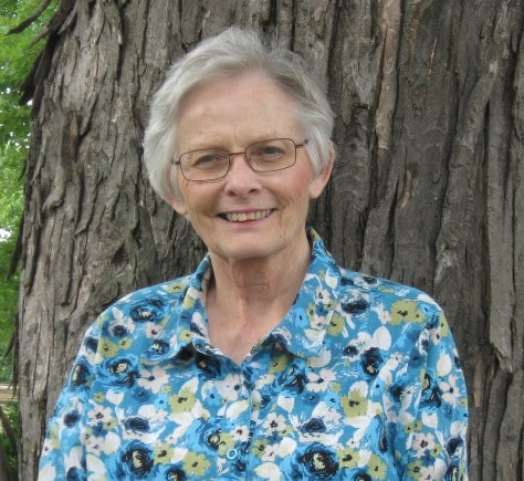 An older adult with short gray hair and glasses, wearing a blue floral shirt, stands in front of a large tree trunk outdoors.